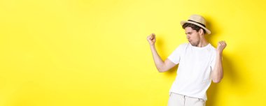 Turizm ve tatil kavramı. Şanslı adam turist seviniyor, yumruk tokuşturuyor ve evet diyor, sarı arka planda mutlu duruyor..