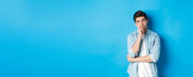 Kızgın yetişkin bir adam gözlerini deviriyor ve sıkılmış görünüyor. Sıradan kıyafetlerle mavi arka plana karşı kayıtsız kalıyor..