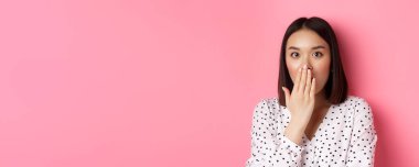 Şok olmuş Asyalı bir kızın görüntüsü. Dedikoducu, nefes nefese, ağzı kapalı, kameraya tamamen güvensizce bakıyor, pembe arka planda duruyor..