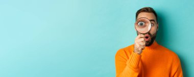 Komik adam büyüteçten bakıyor, bir şeyi arıyor ya da araştırıyor turkuaz arka planda turuncu süveter giyiyor..