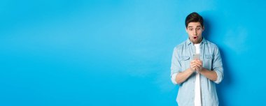 Akıllı telefondan reklamları kontrol ederken şaşkın bakan, telefona bakan, mavi arka planda duran adam..