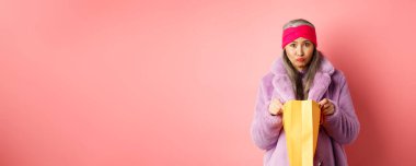 Son sınıf Asyalı üzgün bir kadın alışveriş çantasını açıyor ve pembe arka planda dikilip kameraya üzgün üzgün bakıyor..