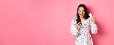 Çekici genç Asyalı kadın online sipariş veriyor, kredi kartı ve cep telefonu tutuyor, internet alışverişi yapıyor, pembe arka planda mutlu duruyor..