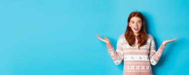 Heyecanlı kızıl saçlı kızın görüntüsü iyi habere tepki veriyor. Şaşırmış görünüyor, ellerini yana doğru açıyor ve gülümsüyor. Kış süveteri içinde mavi arka plana karşı duruyor..