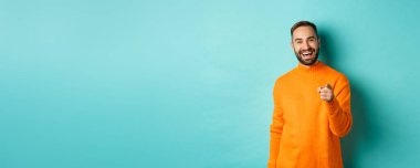 Yakışıklı adam gülüyor ve kamerayı işaret ediyor, onaylamak için başını sallıyor, seninle aynı fikirde, açık mavi arka planda duruyor..