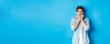 Korkmuş yetişkin bir adam korkunç bir şeye bakıyor, tırnaklarını yiyor ve korkudan titriyor, mavi arka planda duruyor..