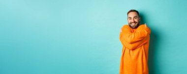 Mutlu adam kendini rahat hissediyor, sıcak bir süveter giyiyor ve kendine sarılıyor, gülümsüyor, açık mavi arka planda duruyor..