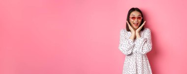Turizm ve yaşam tarzı konsepti. Sevimli güneş gözlüklü ve elbiseli heyecanlı Asyalı kadın, mutluluk çığlığı atıyor ve kameraya bakıyor, pembe arka planda şaşkın şaşkın duruyor..