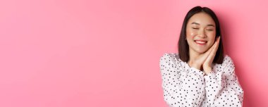 Güzellik ve yaşam tarzı konsepti. Güzel ve rüya gibi Asyalı bir kadının ellerinin üstünde uyuduğu, gözlerini kapadığı ve gülümsediği, pembe arka planda hayal kurduğu yakın plan..