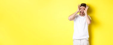 Genç adam komik yüzler ve yapışkan dil gösteriyor, sarı arka planda şakacı duruyor..
