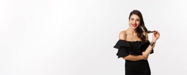 Güzellik ve moda konsepti. Siyah elbiseli, kırmızı dudaklı, saçlarıyla oynayan ve dinleyen, kameraya ilgiyle bakan, beyaz arka planda duran çapkın bir kadın..