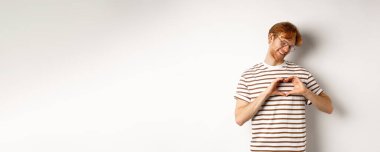 Sevgililer Günü. Kızıl saçlı mutlu erkek arkadaş, gülümseyen ve kalp hareketi gösteren, seni seviyorum, beyaz arka planda duruyorsun..