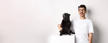 Gözlüklü genç adam köpeğine, hayvan sahibine ve köpeğine sarılıyor sol üst köşedeki reklam teklifine bakıyor, beyaz arka planda duruyor..