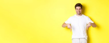 Genç yakışıklı adam işaret ediyor ve aşağı bakıyor, reklam teklifini kontrol ediyor, sarı arka planda duruyor..