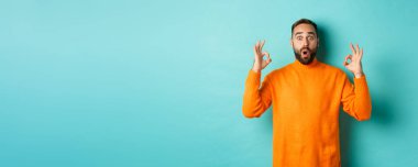 Etkileyici bir adam süper havalı bir şey övüyor, tamam işareti gösteriyor ve kameraya bakıyor, açık mavi arka plan.