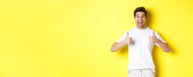 Heyecanlı yakışıklı adam başparmağını kaldırıyor, onaylıyor ve evet diyor, sarı arka planda duruyor..