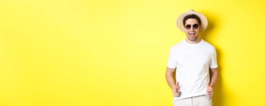 Tatilde seninle flört eden, kamerayı işaret eden ve göz kırpan güneş gözlüklü, sarı arka planda yaz şapkası takan kendinden emin ve küstah bir adam..