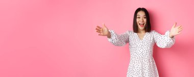 Mutlu Asyalı kız ellerini kameraya uzatıyor, sarılmaya uzanıyor, gülümsüyor ve bir şeyi tutmak istiyor, pembe arka planda duruyor..