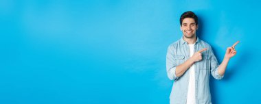Çekici yetişkin bir adamın portresi gülümsüyor, parmaklarını logoya ya ya da afişe doğrultuyor, mavi arka plana karşı reklam gösteriyor..