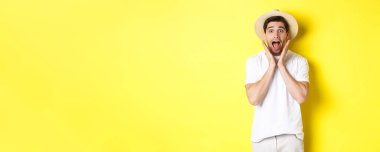 Turizm ve yaz kavramı. Hasır şapkalı şaşırmış erkek model, sarı arka planda duran, özel tekliflere şaşırmış görünüyor..