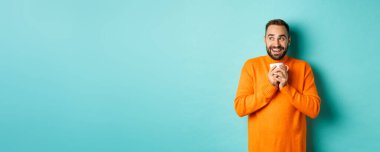 Mutlu genç adam kahve içiyor, gülümsüyor ve rüya gibi bir yüzle soluna bakıyor, turkuaz arka planda dikiliyor..