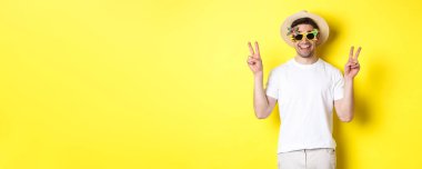 Turizm ve yaşam tarzı kavramı. Mutlu adam seyahatten zevk alıyor, yaz şapkası ve güneş gözlüğü takıyor, fotoğraf için barış işaretleriyle poz veriyor, sarı arka plan.