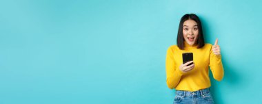 E-ticaret ve online alışveriş konsepti. Akıllı telefon uygulaması, tavsiye cihazı, mavi arka planda duran Asyalı kadın, heyecanlı ve şaşırmış..