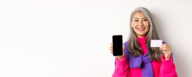 İnternetten alışveriş. Gülümseyen Asyalı son sınıf öğrencisi kadının boş cep telefonu ekranı ve plastik kredi kartı göstermesi, mutlu görünmesi, beyaz arka planda durması..