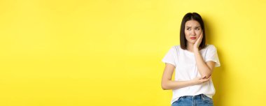 Üzgün Asyalı kadın yanağa dokunuyor ve kaşlarını çatıyor, diş ağrısı çekiyor, dişçiye görünmesi gerekiyor, sarı arka plana karşı kasvetli duruyor..