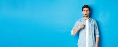 Gergin adam kendini işaret ediyor ve kafası karışık görünüyor. Mavi arka plan üzerinde sıradan kıyafetler giyiyor..