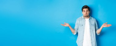 Kafası karışmış genç adam omuzlarını silkiyor, hiçbir şey bilmiyor, habersiz görünüyor, mavi arka planda duruyor..