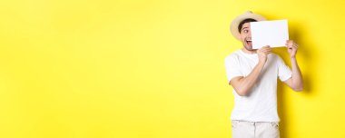 Tatildeki heyecanlı adam logonuz için boş bir kağıt parçası gösteriyor, yüzünde tabela tutuyor ve gülümsüyor, sarı arka planda duruyor..