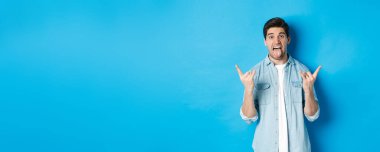Heyecanlı yakışıklı adam eğleniyor, rock 'n' roll işareti yapıyor ve dili yapışkan, mavi arka planda duruyor..