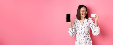 Mutlu Asyalı genç kadın internet alışverişi, akıllı telefon ekranı gösteriyor ve kredi kartından memnun görünüyor, pembe arka planda duruyor..