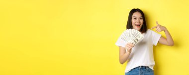 Gülümseyen Asyalı kadın parayı işaret ediyor, dolar gösteriyor, sarı arka planda duruyor..