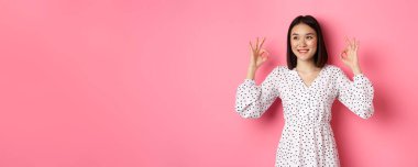 Hoş, genç Asyalı bir kadın iyi bir işaret veriyor, övgüler yağdırıyor ve onaylıyor, tatmin olmuş görünüyor, pembe arka plana karşı duruyor..