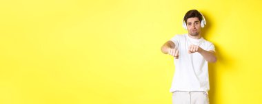 Kulaklıkla müzik dinleyen, dans eden, beyaz elbiseli, sarı stüdyo arka planında duran havalı adam..