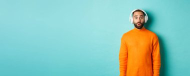 Kulaklıkla müzik dinleyen, sesten etkilenmiş kameraya bakan, turkuaz arka planda duran yetişkin bir adam..