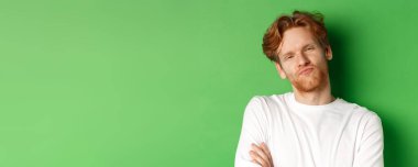 Duygular ve moda konsepti. Kızıl saçlı, sakallı, büzüşmüş dudakları olan, kameraya bakan, yeşil arka planda duran şüpheci genç adamın yakın çekimi..