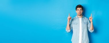 Hoşnutsuz ve şüpheci erkek modelin portresi parmakları havada, nahoş bir şeye bakıyor, mavi arka planda duruyor..