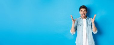 Hüsrana uğramış genç adam bağırıyor, gergin görünüyor, el sıkışıyor, mavi arka planda duruyor..