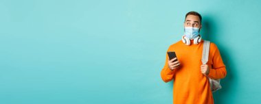 Yüz maskeli genç adam cep telefonu kullanıyor, sırt çantasını tutuyor, sola şaşkın şaşkın bakıyor, açık mavi arka planda duruyor..