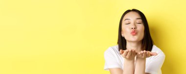 Sevgililer Günü konsepti. Güzel Asyalı bir kadının dudaklarını büzüştürmesi ve el ele tutuşması, kameraya kapalı gözlerle hava öpücüğü üflemesi, sarı arka planda durması..