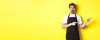 Siyah önlüklü şaşkın garson, tanıtım teklifini gösteriyor, parmaklarını sağa gösteriyor ve şaşırmış görünüyor, sarı arka planda üniformalı duruyor..