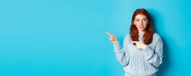 Süveter giymiş güzel kızıl saçlı bir kız resmi, parmaklarını logoya gösteriyor, garip bir şekilde gülümsüyor, mavi arka planda duruyor..