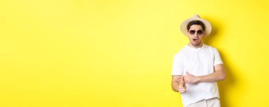 Yakışıklı adam parmak tabancasıyla ateş ediyor ve şımarık görünüyor. Yaz tatili için giyinmiş, hasır şapka ve güneş gözlüğü takıyor. Sarı arka planda duruyor..