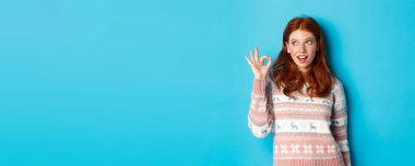 Süveter giyen aptal kızıl kadın, tamam işareti gösteriyor ve sol üst köşedeki logoya bakıyor, mavi arka planın üzerinde duruyor..