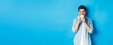 Genç adam mutlu bir yüzle kahve içiyor, mavi arka planda duruyor..