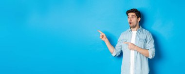 Şaşırmış ve şaşırmış bir adam terfiye bakıyor, parmaklarını reklamlara gösteriyor, mavi arka planda duruyor..