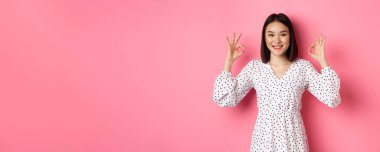Hoş, genç Asyalı bir kadın iyi bir işaret veriyor, övgüler yağdırıyor ve onaylıyor, tatmin olmuş görünüyor, pembe arka plana karşı duruyor..